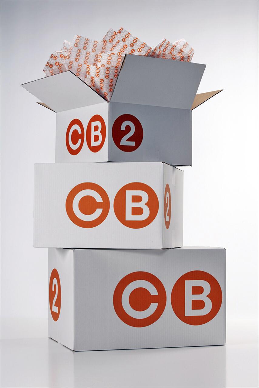 Stack of three white cardboard boxes with orange "CB2" logos, one box open with packing material inside.
