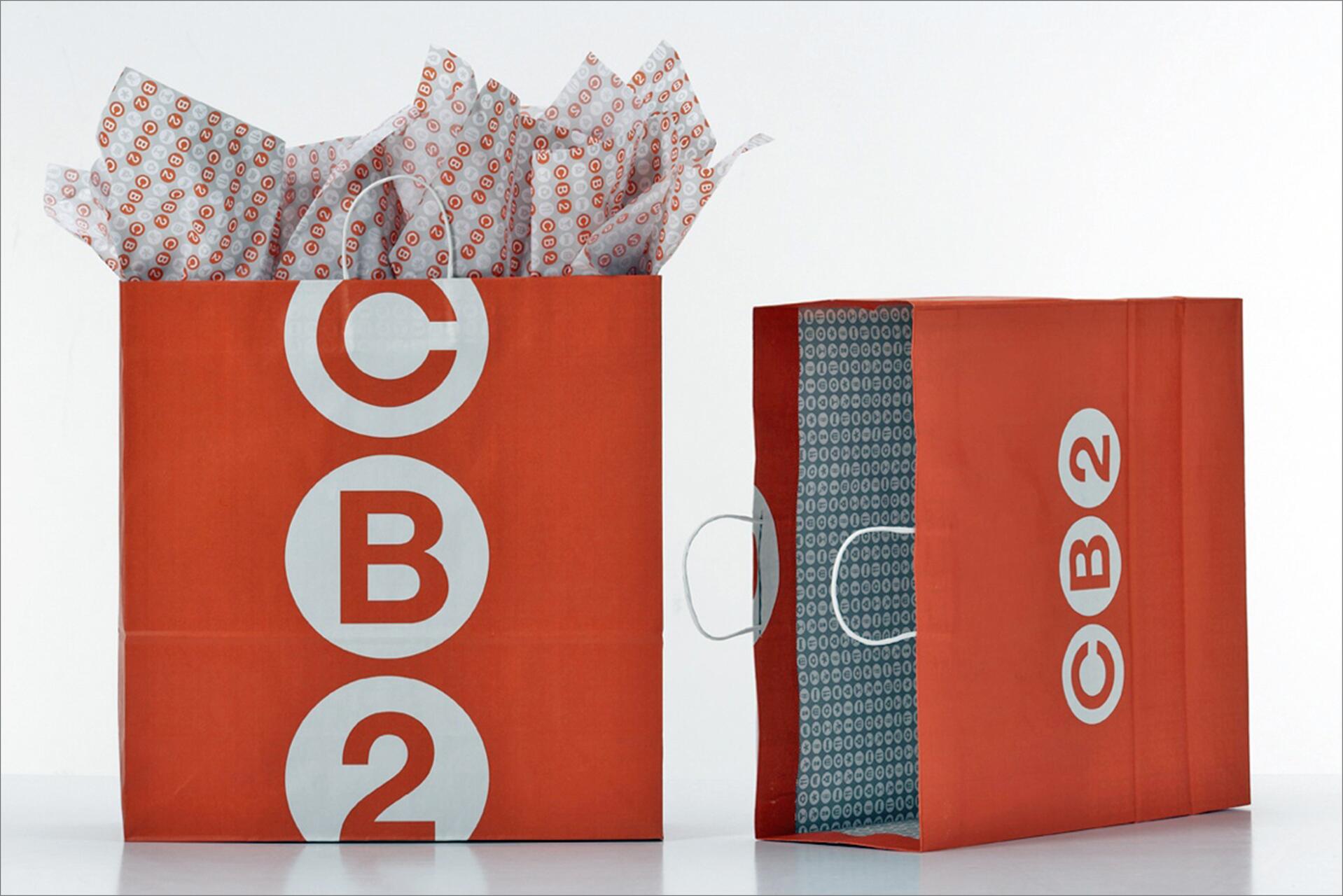 Two red shopping bags with white "CB2" logos. The bag on the left is filled with tissue paper, while the right bag is empty, displaying white handles and a patterned interior.