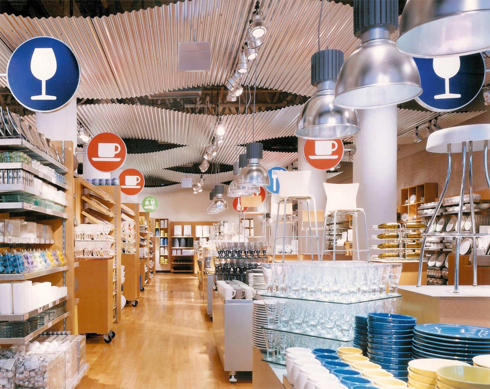 Store interior with wooden shelves and racks displaying dishes, glasses, and kitchenware. Hanging signs indicate sections with icons of wine glasses and teacups. Metal light fixtures hang from a wavy ceiling. Bright and organized layout.