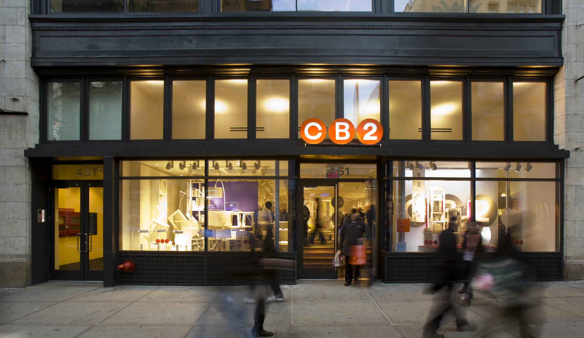 Storefront of a CB2 store with large glass windows, warm interior lighting, and a bright orange sign. Blurred pedestrians walk by on the sidewalk, suggesting motion.