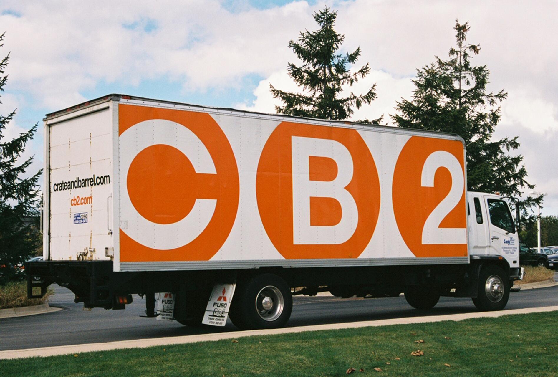 Delivery truck with a white and orange CB2 logo parked near pine trees under a partly cloudy sky.