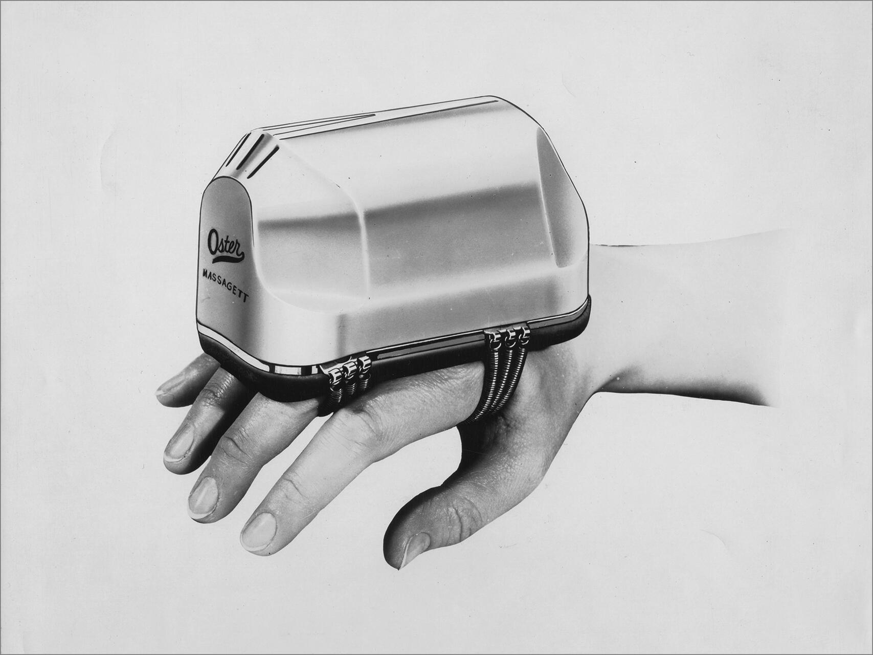 A vintage hand massager labeled "Oster MASSAGETT" worn on a hand with fingers visible below the device, against a plain background.