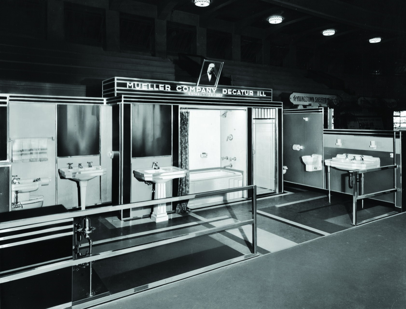 Black and white photo of a mid-20th century bathroom showroom display featuring sinks, a bathtub, a shower, and toilets. The setup is organized in a row under signage for a plumbing company.
