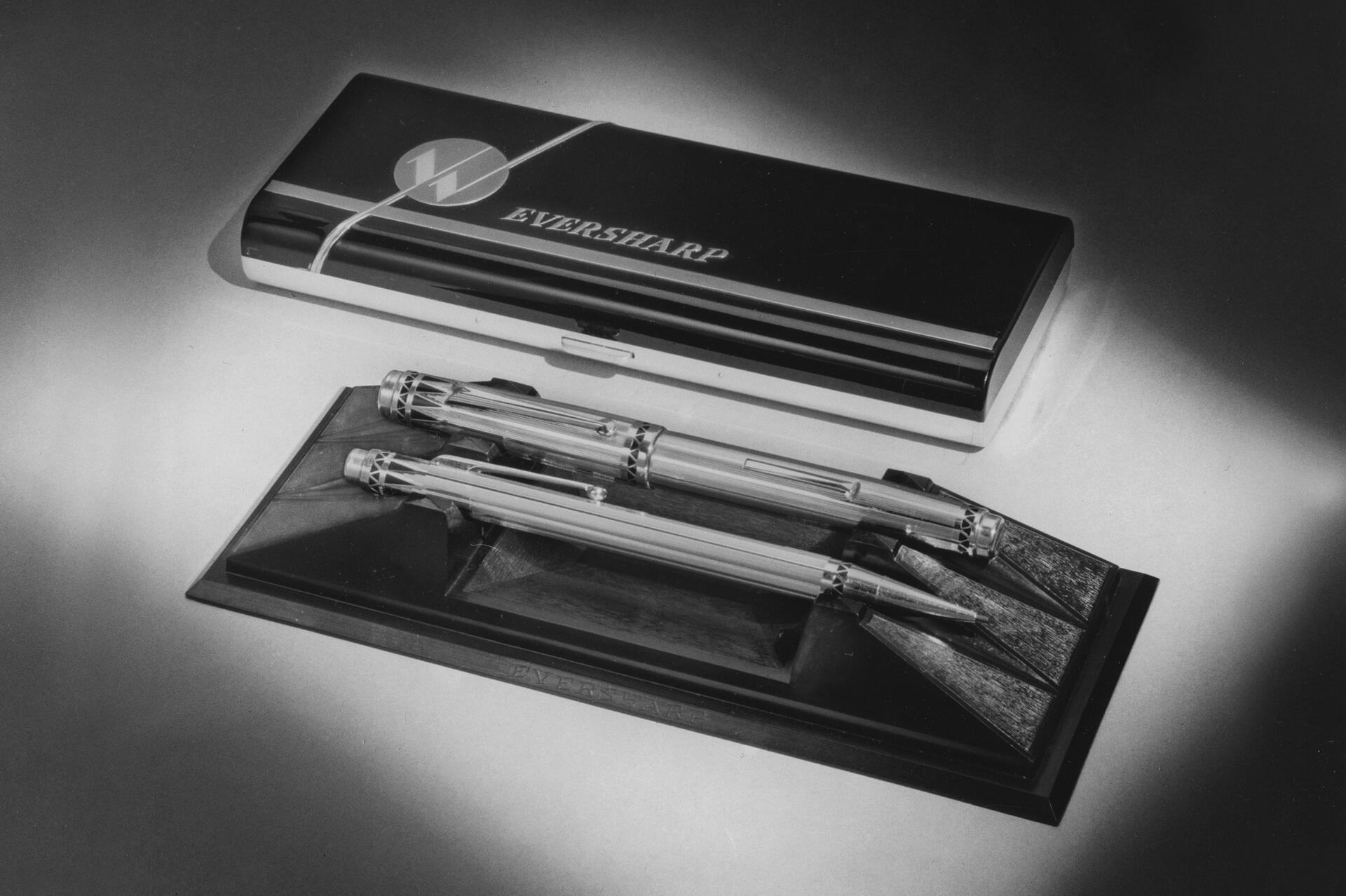 Black and white photograph of two metal pens, displayed on a stand, with a sleek, closed case labeled "Eversharp" beside them.