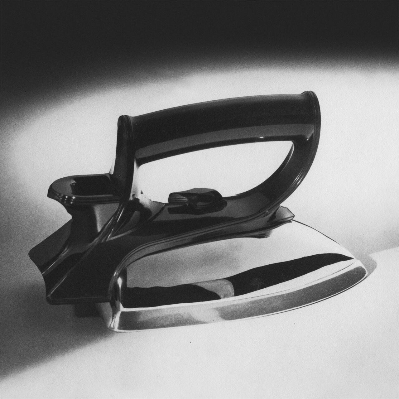 Vintage black and white photograph of a classic metal steam iron with a sleek handle, casting a shadow on a light surface.