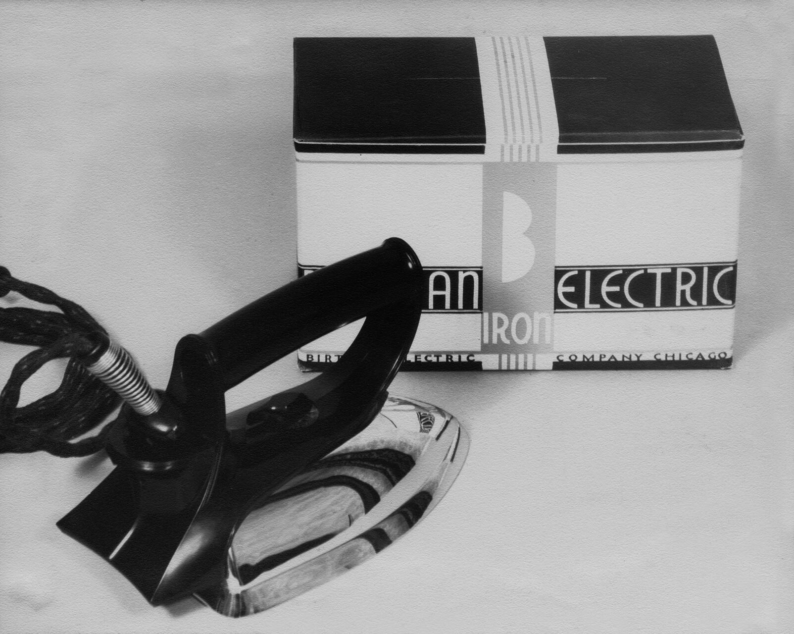 A vintage black electric iron with a twisted cord sits next to its box, featuring bold, retro lettering and design.