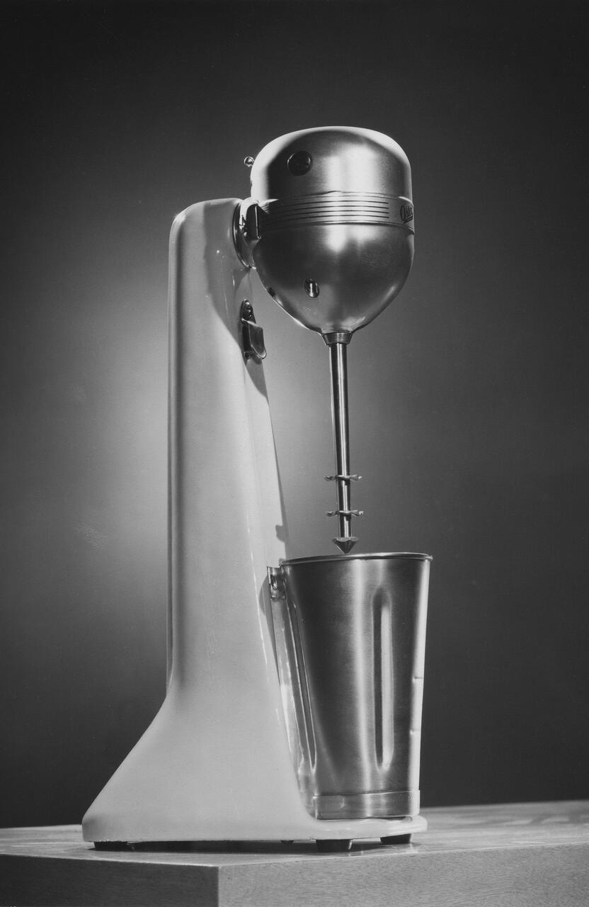 Vintage black and white photograph of a standing electric mixer with a stainless steel cup and a sleek, rounded design.