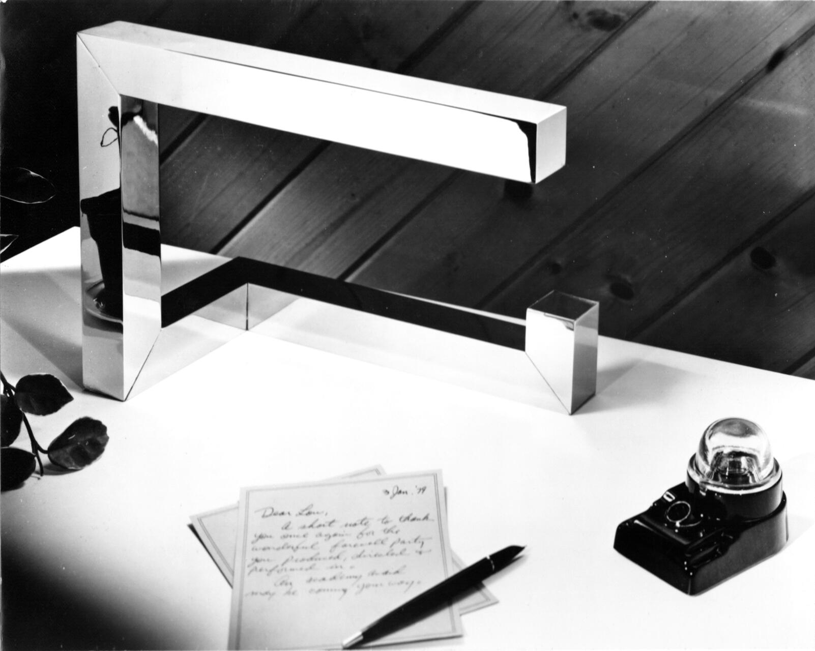 A modern, chrome sculpture resembling a large staple sits on a white table. Nearby are a few leaves, a black pen, a handwritten note, and an old-fashioned black telephone with a clear rotary dial. Wood panel wall in the background.