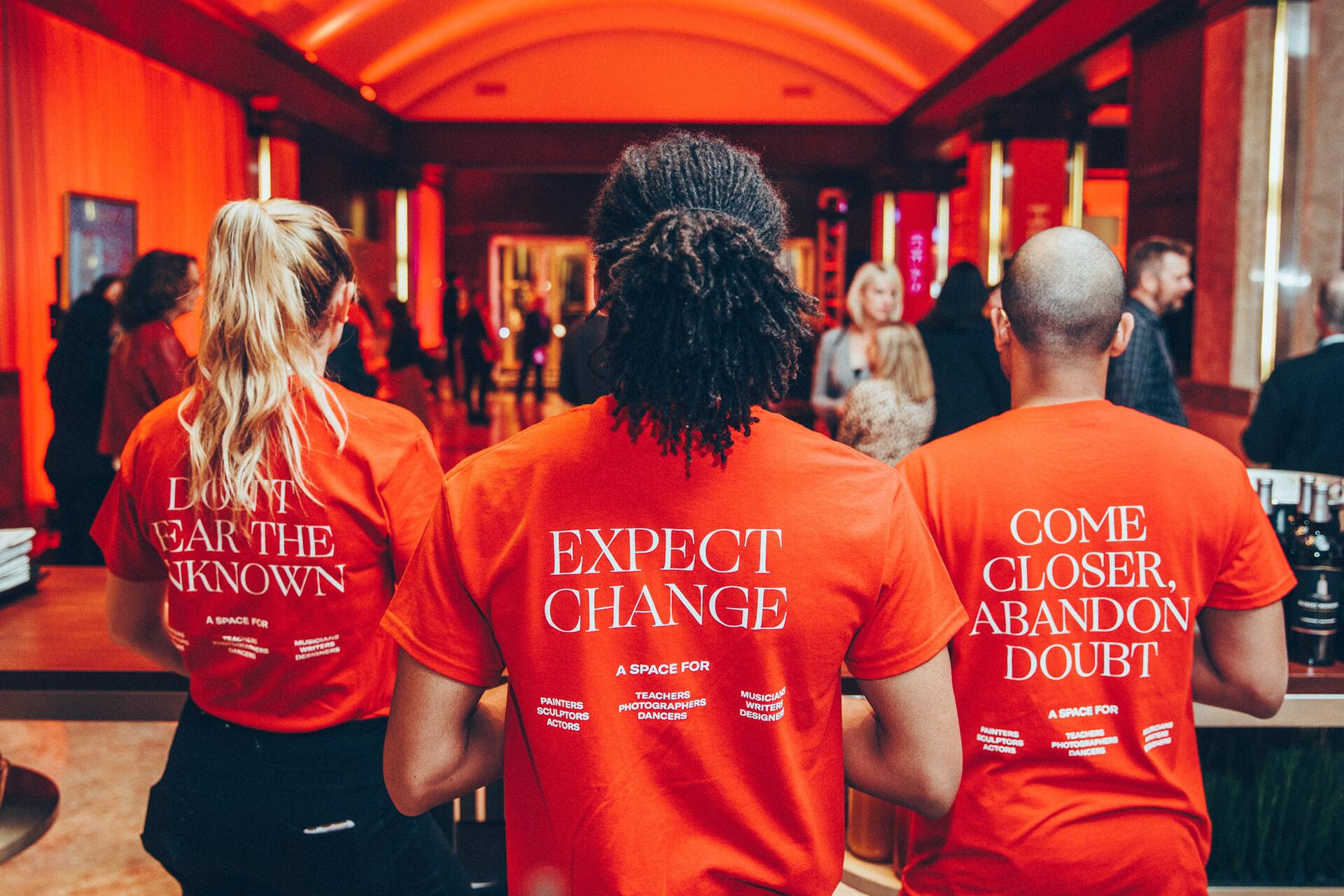 Three people with their backs to the camera wearing red shirts with motivational text, inside a warmly-lit venue with a crowd in the background.