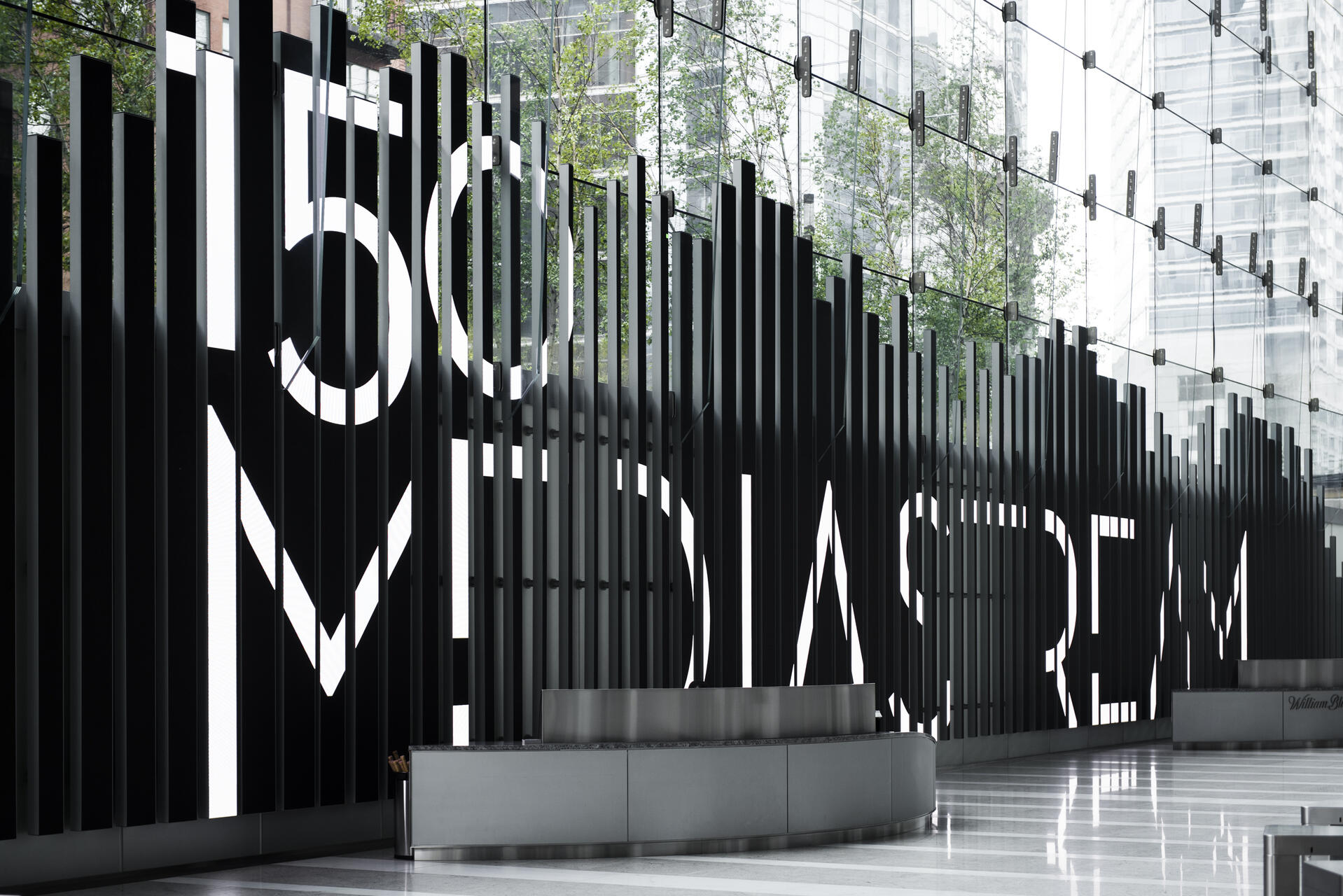 Modern atrium with tall black vertical bars spelling "150 Mediastream" in bold white letters, with glass walls reflecting trees and city buildings outside.
