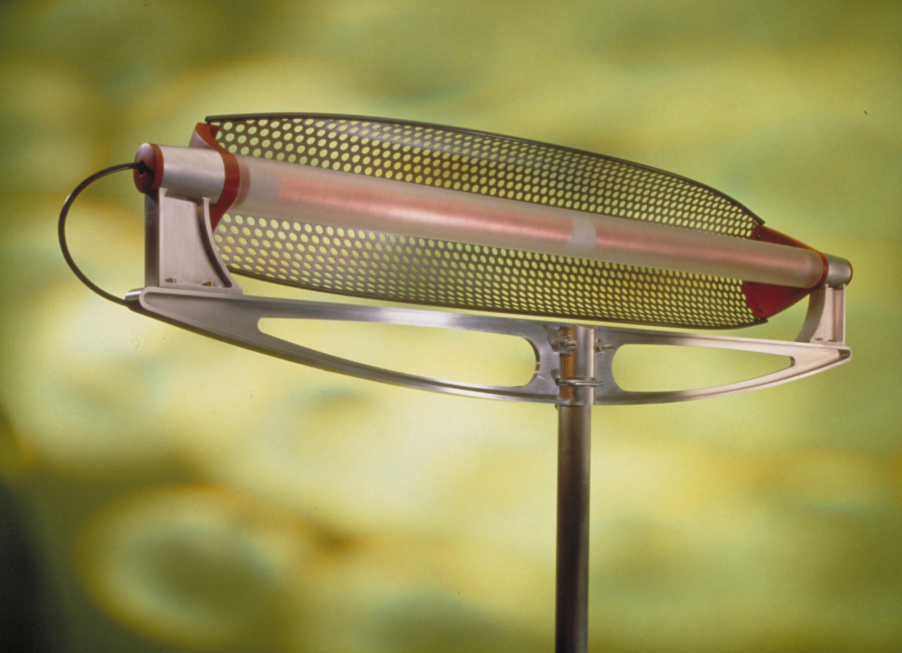 Perforated metal sculpture with a cylindrical rod on a stand against a blurred yellow-green background.