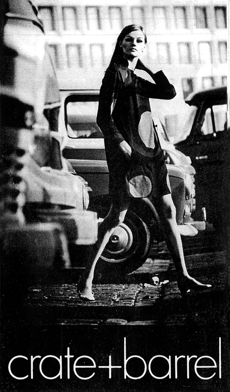 Black and white photo of a woman in a patterned dress walking confidently between parked cars, with a building blurred in the background. The Crate & Barrel logo is at the bottom.