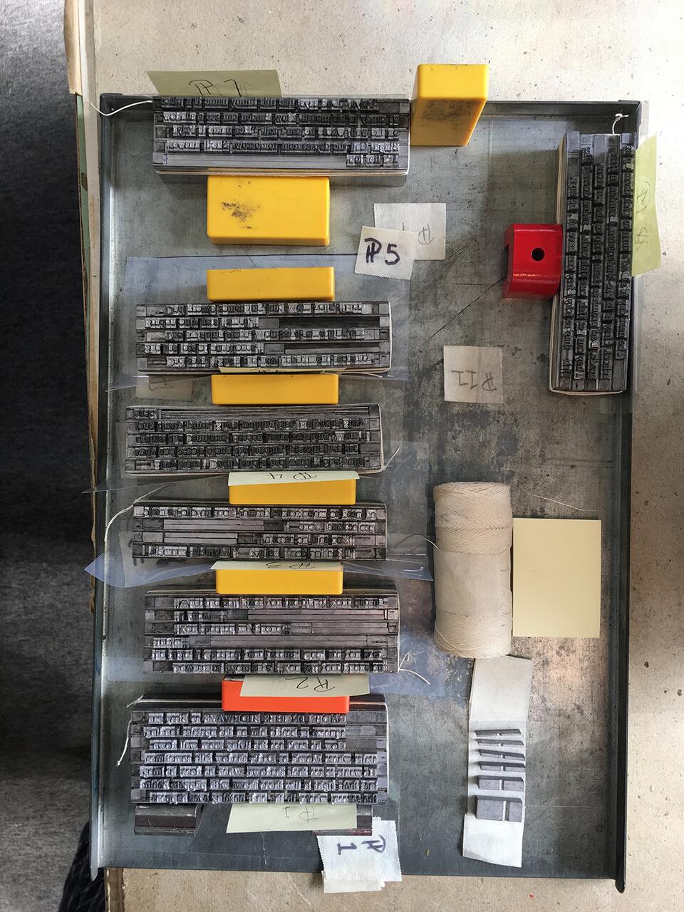 Metal typesetting blocks organized in rows with yellow and red separators on a metal tray. Labels with numbers are attached to some blocks. A roll of string and yellow note card are nearby. Shadow falls on part of the tray.