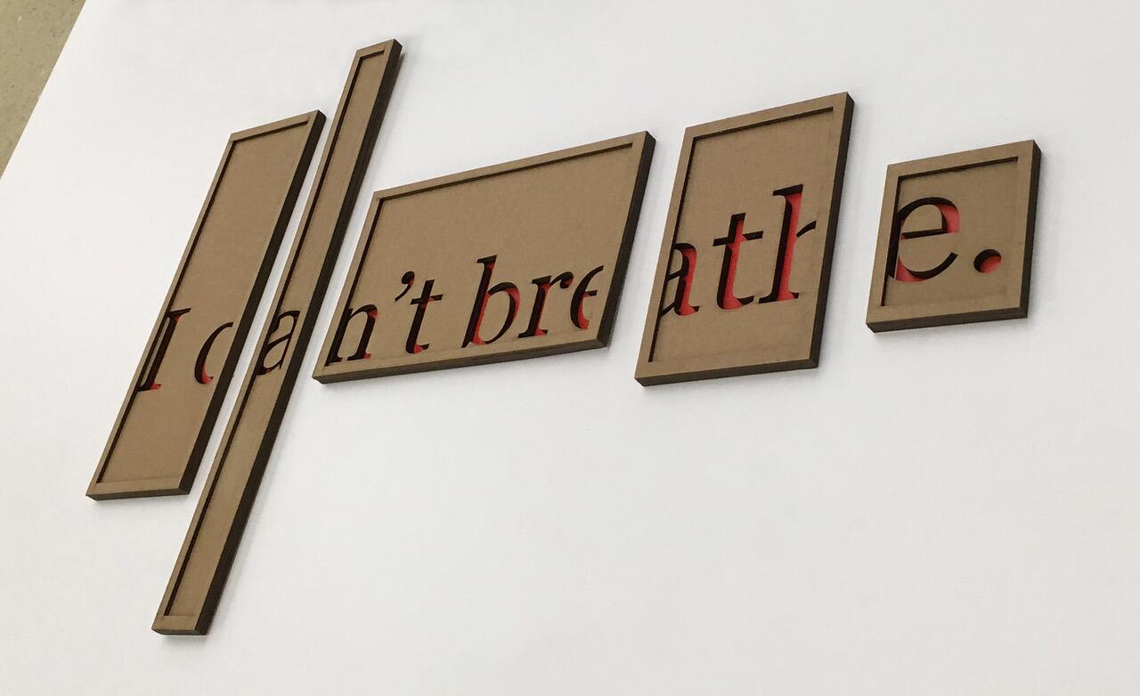 Brown framed cutouts spell "I can't breathe" on a white wall, with red accents visible through the lettering.
