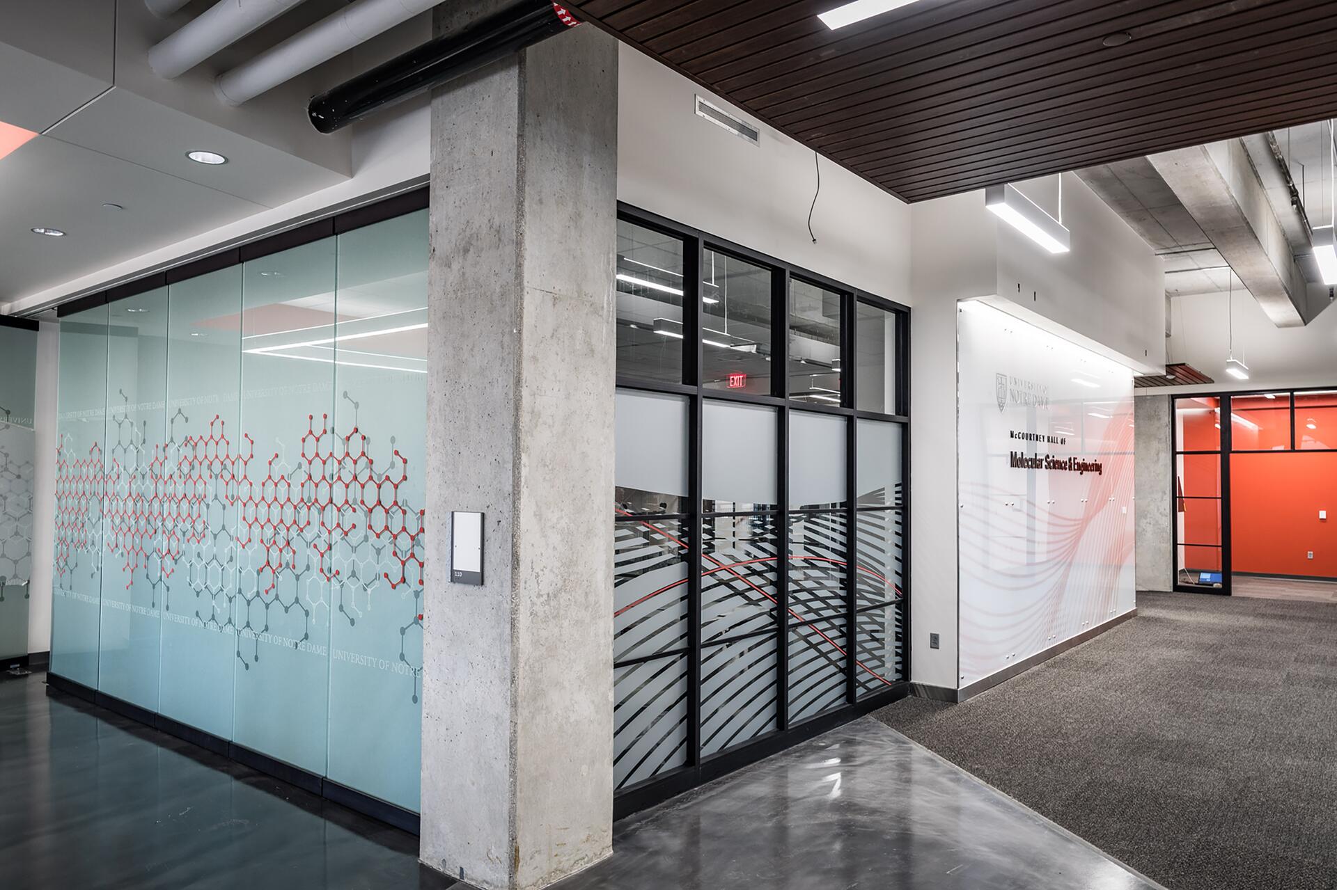 Modern hallway with glass walls and molecular design patterns. Concrete pillar and white walls with black-framed windows. Bright overhead lighting, polished floors, and a red accent wall in a visible room.