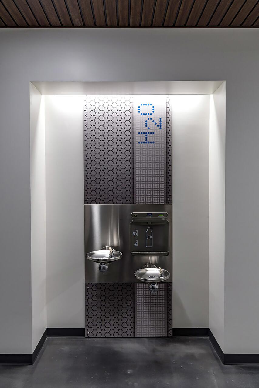 Wall-mounted stainless steel water fountain and bottle filling station with geometric patterned tiles, illuminated by overhead lighting.