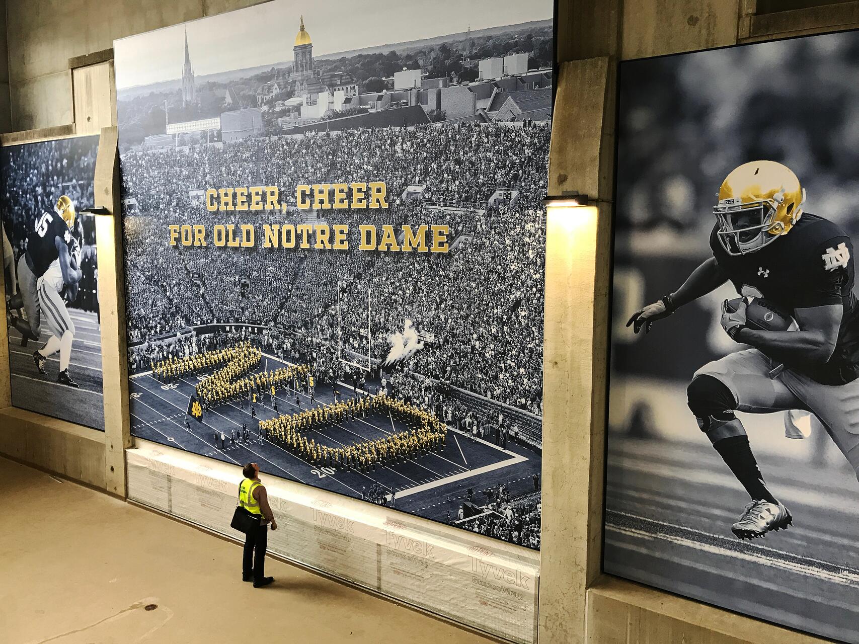 Large stadium mural featuring a football game with marching band, flanked by images of football players. A person in a safety vest looks up at the mural.