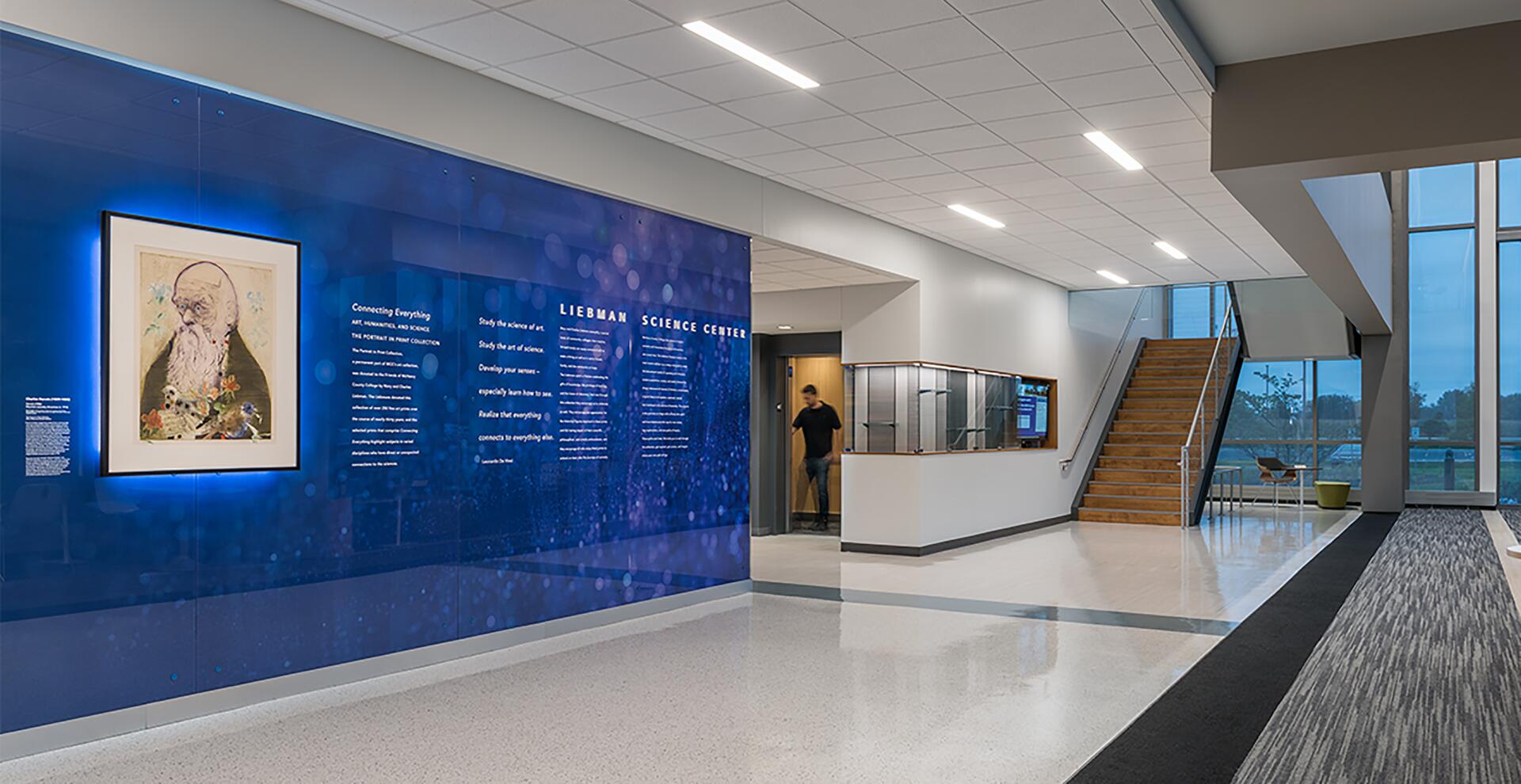 Modern lobby with a blue illuminated wall featuring text and artwork, labeled "Liebman Science Center." A person exits a doorway near a staircase leading to a window overlooking a green landscape.