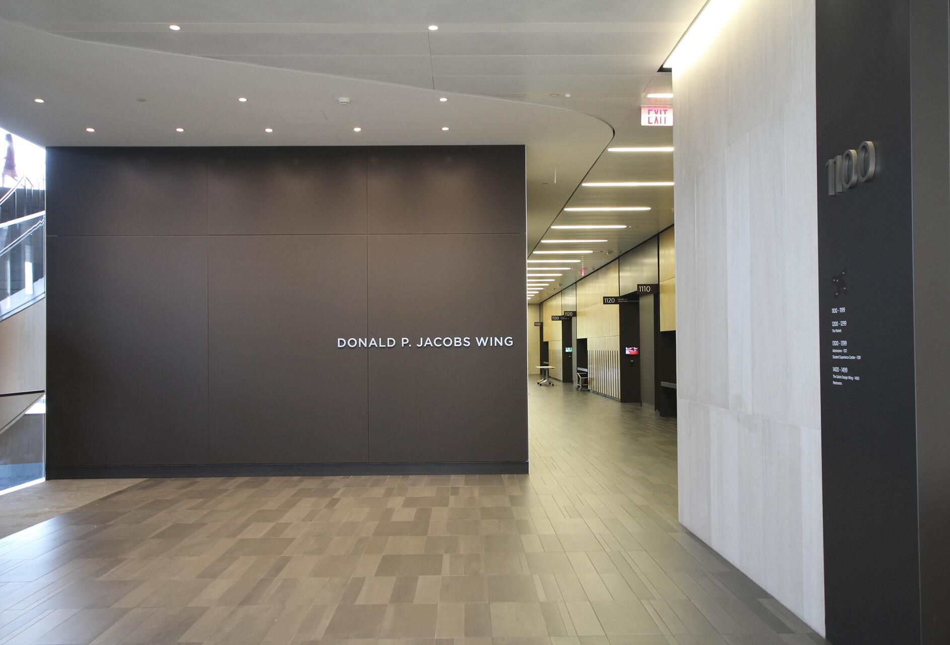 Modern hallway with gray walls, labeled "Donald P. Jacobs Wing." The corridor is brightly lit, with offices and rooms numbered along the right side. Sleek design with dark floor tiles and overhead lights.