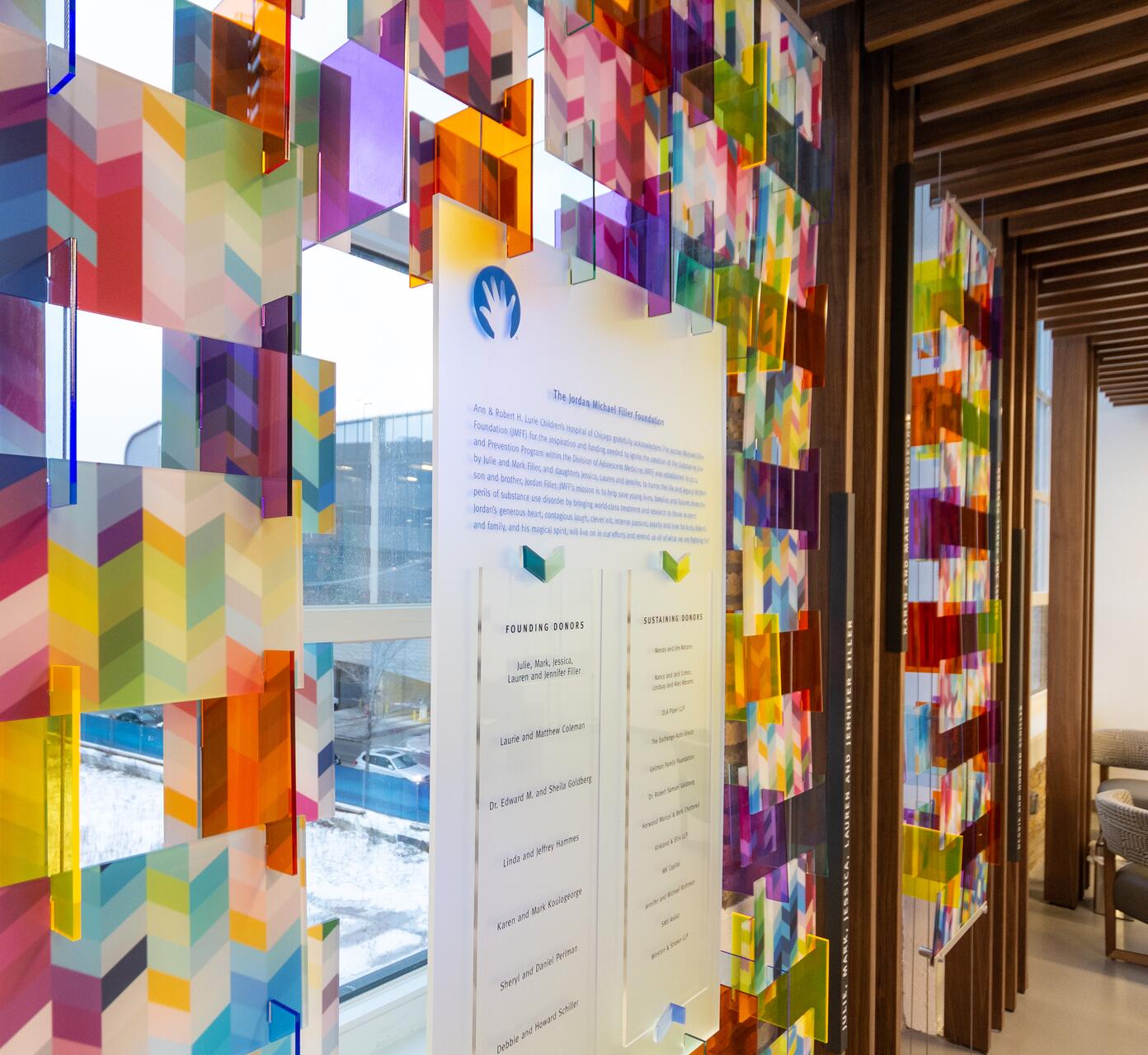Colorful geometric panels with chevron patterns hang in a wooden-framed corridor next to a glass panel listing donors. Natural light filters through a window, highlighting the vibrant colors.