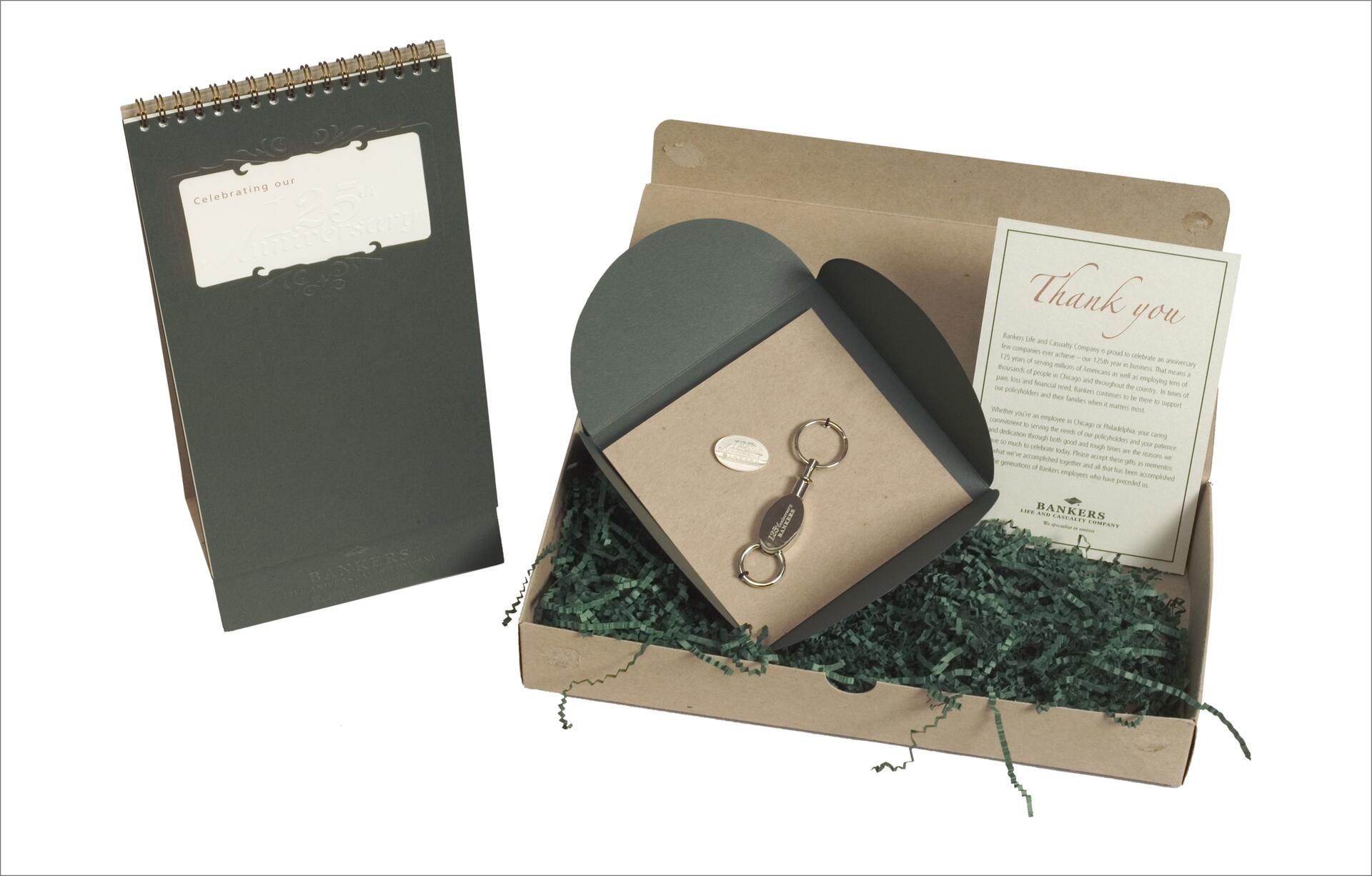 A spiral-bound desk calendar and a gift box with shredded green paper. The box contains a keychain and a thank-you card. The calendar cover features embossed text celebrating a 25th anniversary.