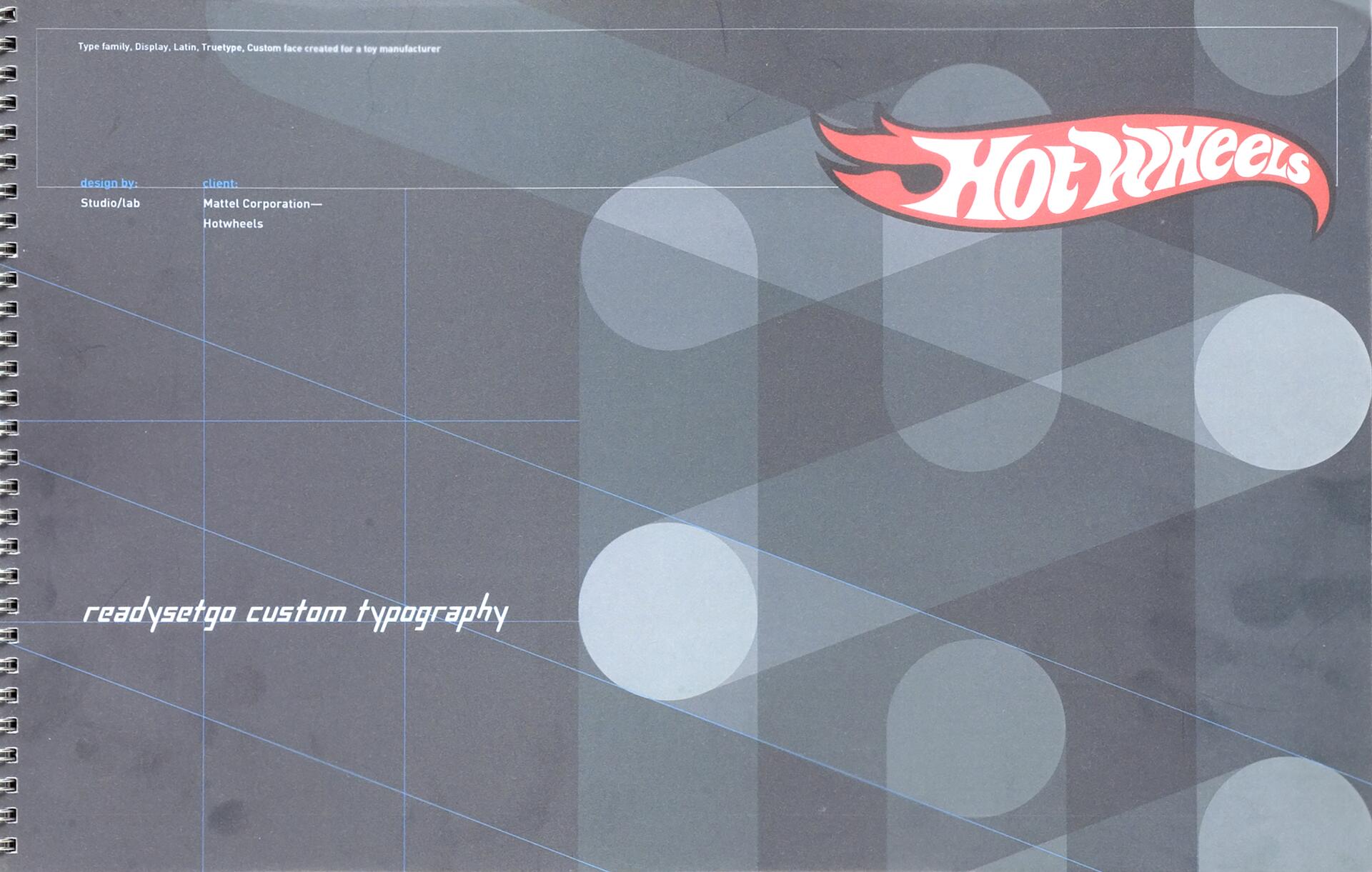 Spiral notebook cover with a geometric design in shades of gray and blue, featuring the "Hot Wheels" logo in red and white at the top right. Text reads "readysetgo custom typography."