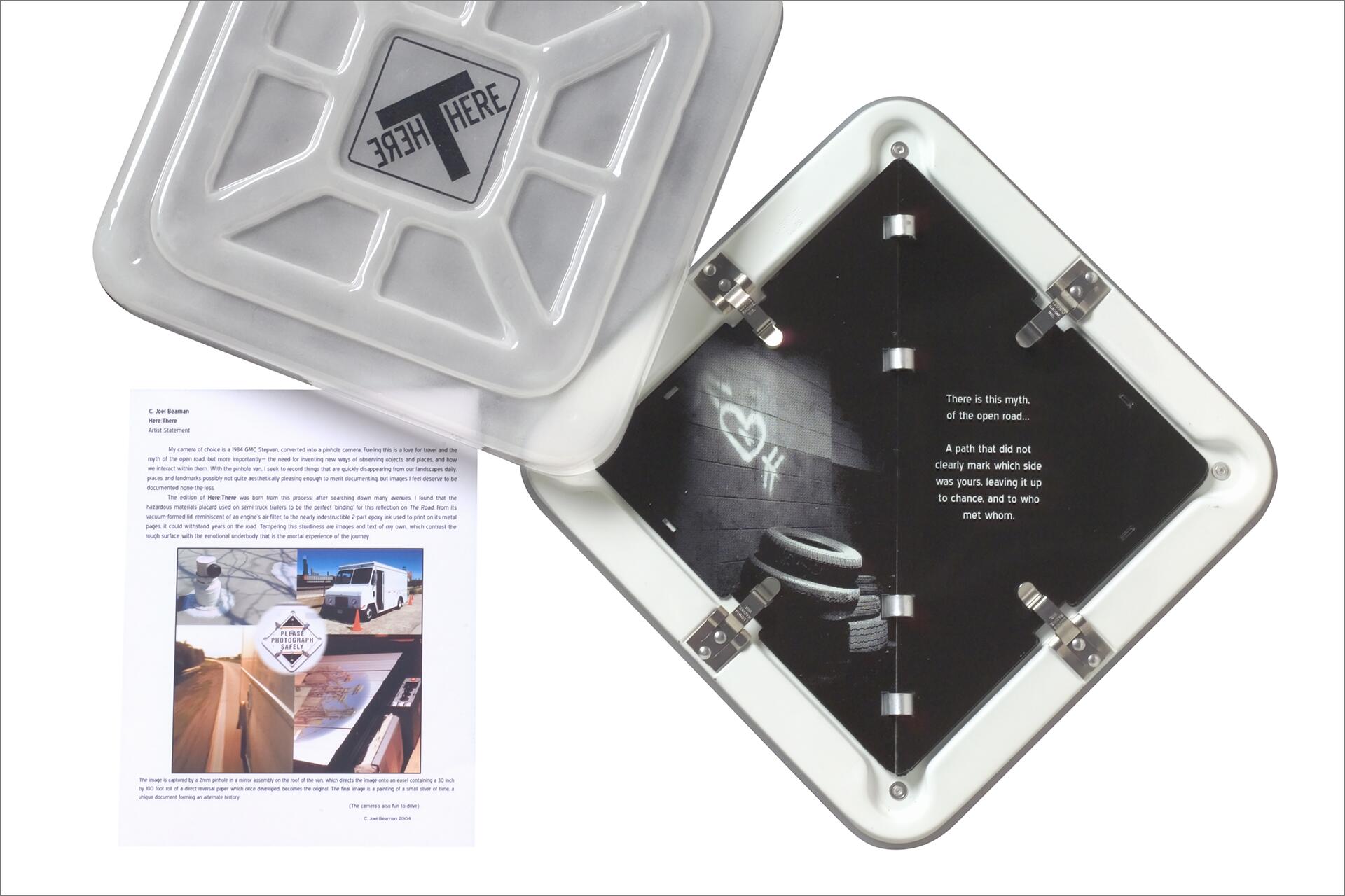 Open gray container with "Here There" logo and a booklet inside, featuring a photo of a tire stack against a wall and text about an open road. Nearby, a printed artist statement with smaller images, including a truck and landscapes.
