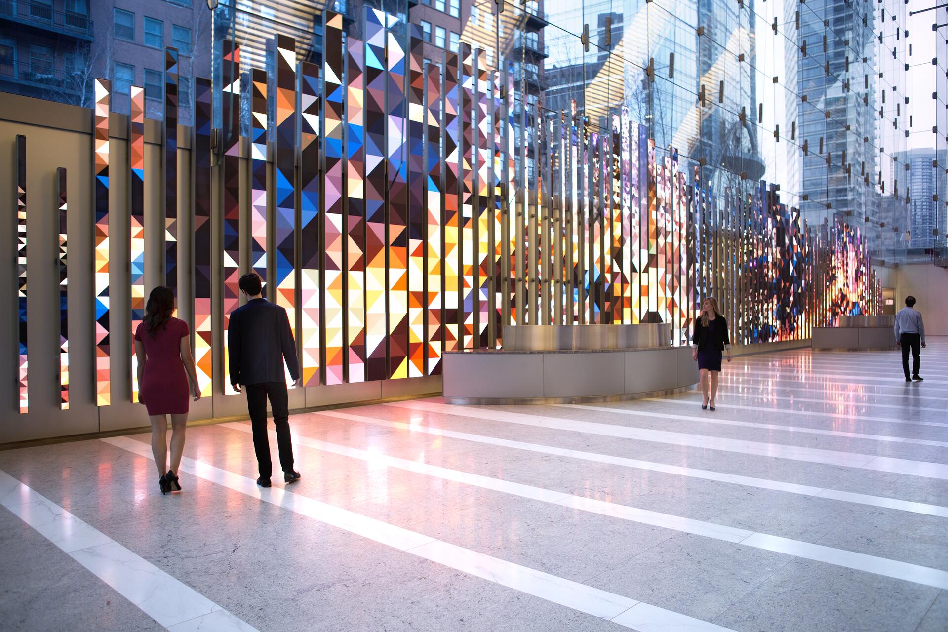 Colorful geometric wall art in a modern building lobby, reflecting vibrant patterns on the glossy floor. People walk along the illuminated corridor, with urban skyscrapers visible through glass windows.
