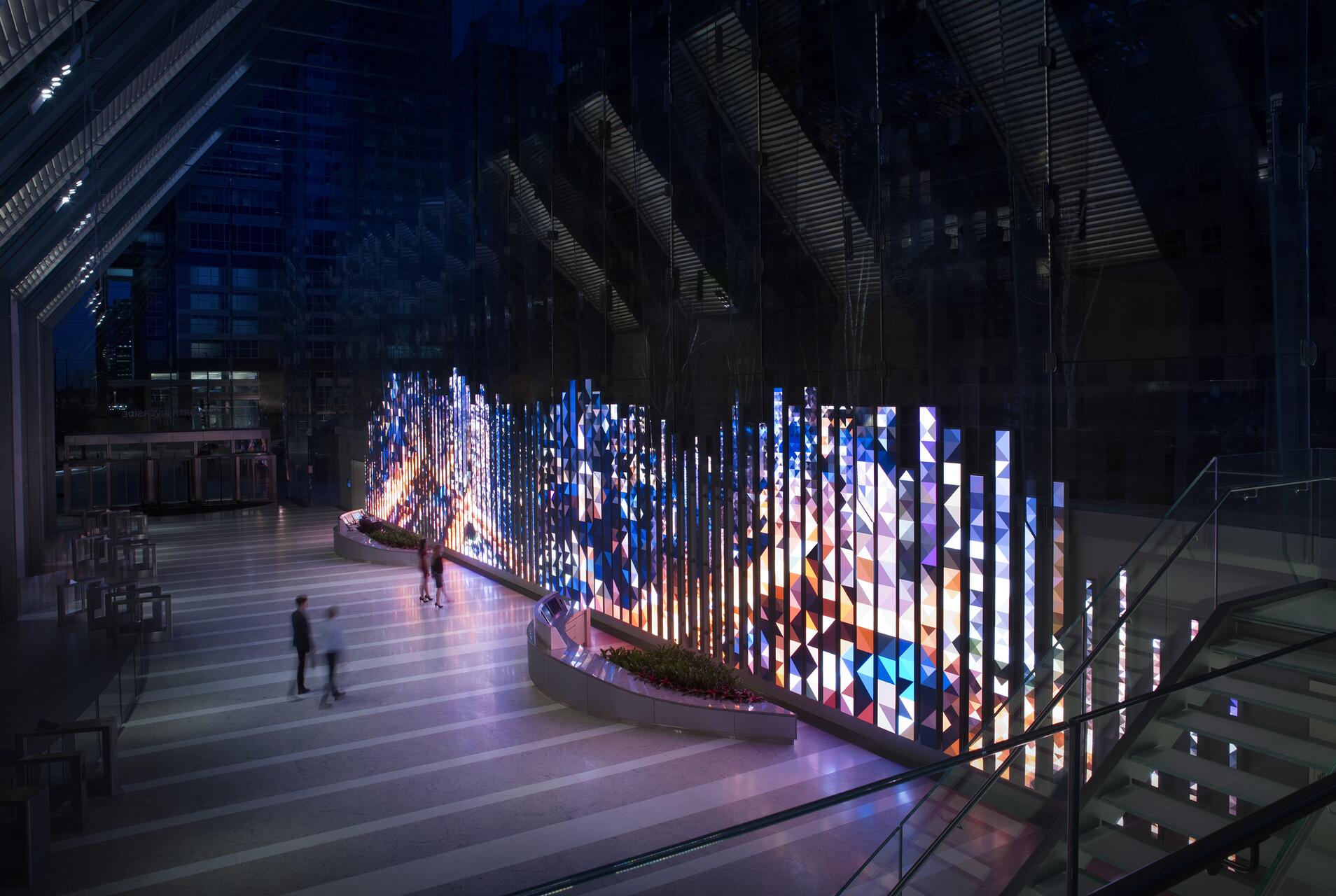Geometric light installation in a modern building lobby, featuring vibrant, illuminated columns of various heights with a colorful triangular pattern. Two people walk by on a striped floor, surrounded by glass walls reflecting the artwork.
