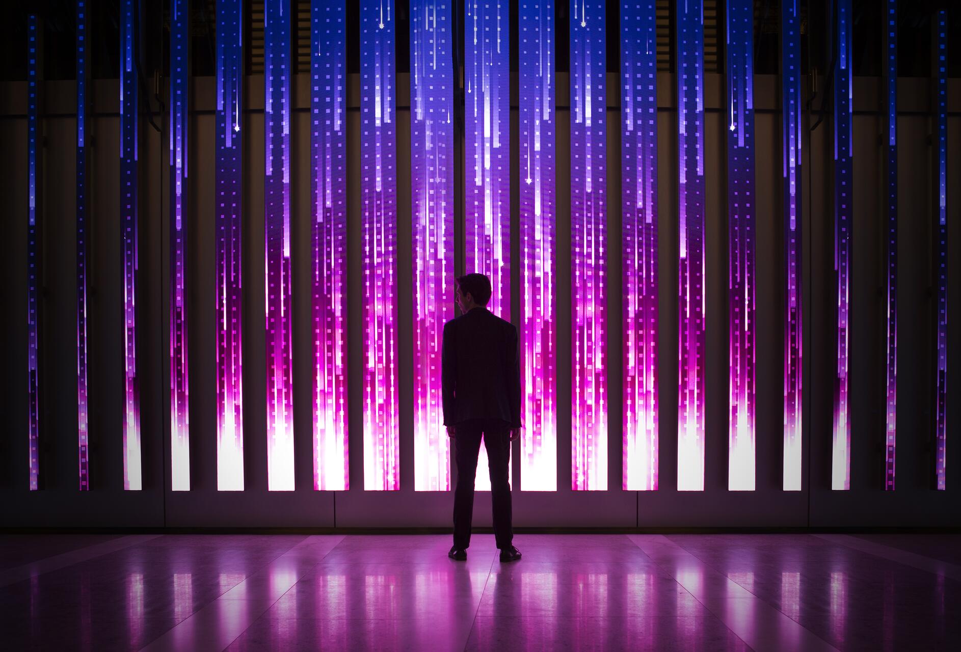 Silhouette of a person facing an illuminated wall with vertical blue and pink light strips, creating a futuristic, digital atmosphere.