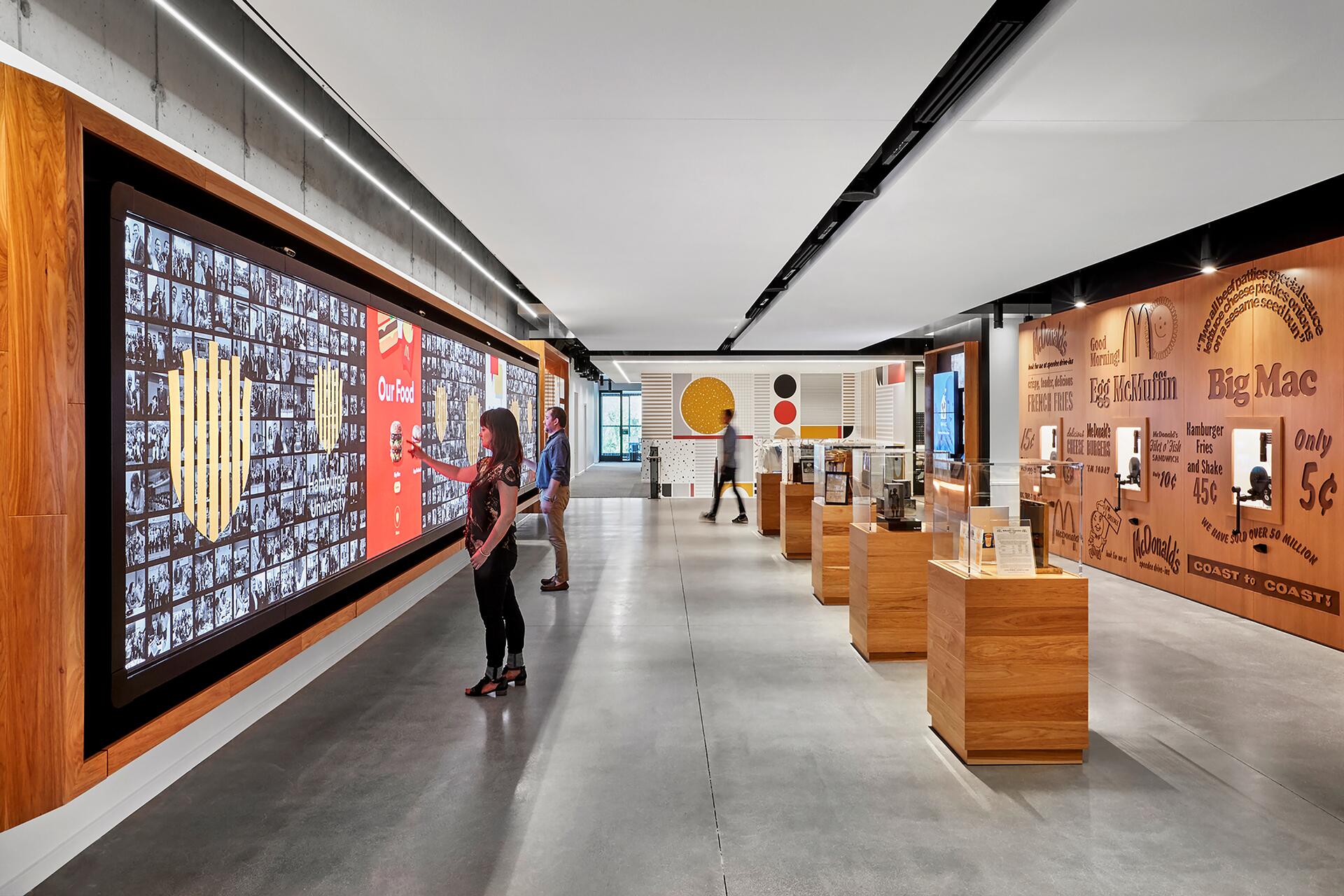 Modern museum exhibit with interactive displays and wall graphics. Visitors engage with large touchscreens displaying historical images and captions. On the right, wooden display cases showcase artifacts, while walls feature vintage advertisements and logos.