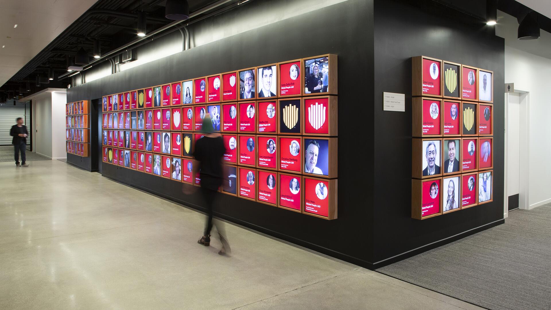 A hallway with illuminated photo displays on a wall, featuring various portraits against a red background. Two people walk past, the floor is polished concrete, and the ceiling has exposed pipes.