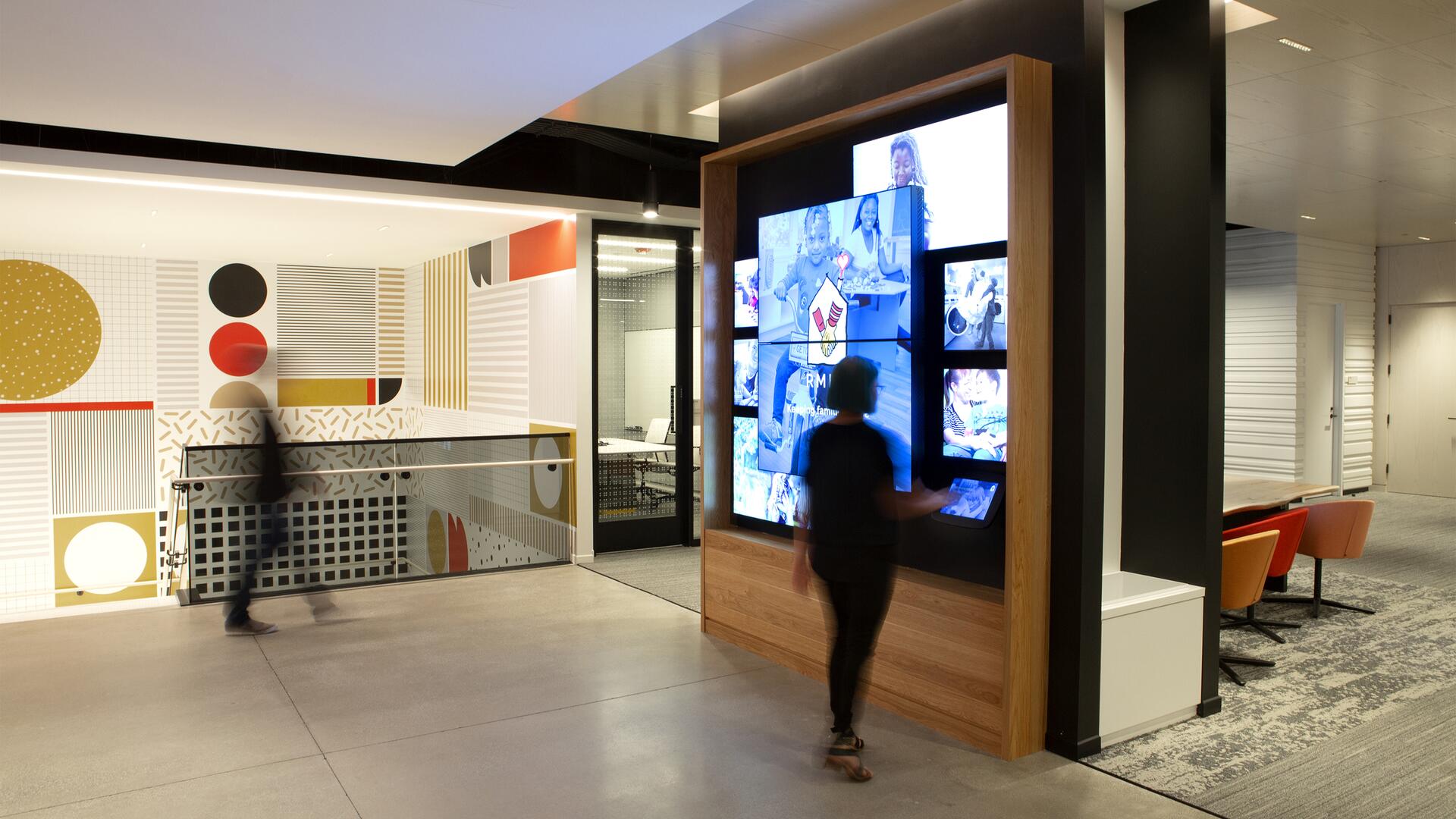 Modern office hallway with colorful geometric wall patterns. A digital display shows multiple images. A person interacts with the screen, while another walks nearby. Bright lighting and sleek design feature prominently.