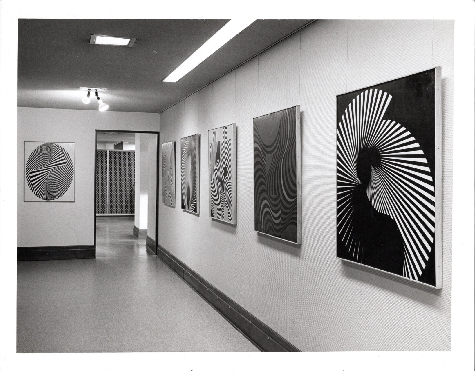 A gallery hallway with black and white, spiral-patterned abstract artwork on the walls. The pieces create optical illusions with wavy and circular lines. Overhead lights illuminate the space.