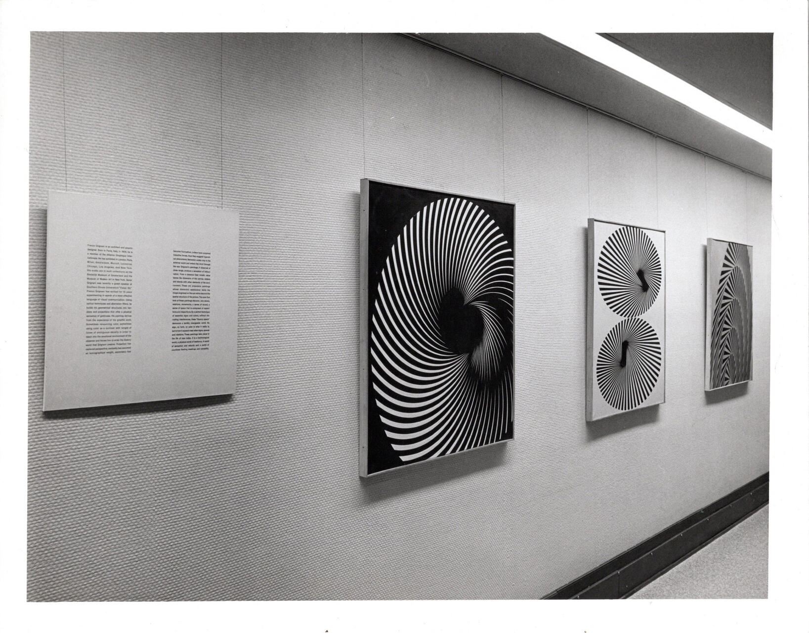 Black and white photograph of a gallery wall displaying four artworks. Three feature swirling, hypnotic patterns of concentric lines, while one has small text blocks. The setting is a minimalist, contemporary space with a smooth wall texture and overhead lighting.