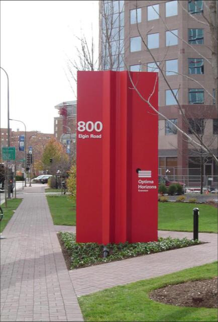 Red monument-style sign with "800 Elgin Road" and "Optima Horizons" on it. Positioned on a sidewalk with grassy areas and surrounded by buildings in an urban setting.