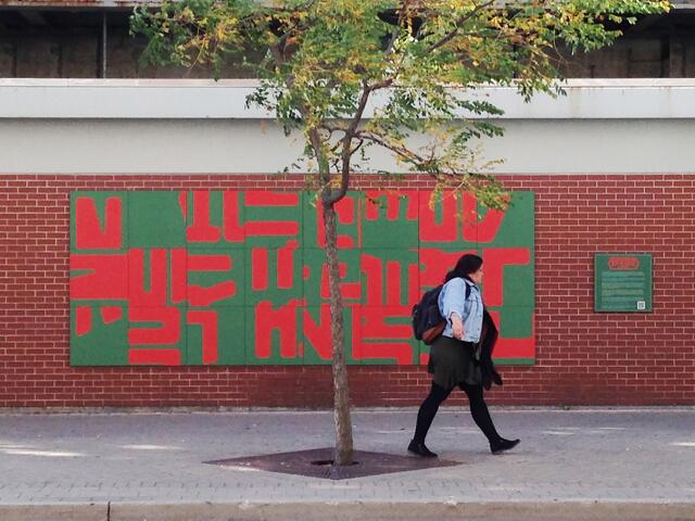 A person walks past a wall mural with red and green abstract patterns, next to a small tree. The wall is brick, and the setting is a sidewalk in a city.