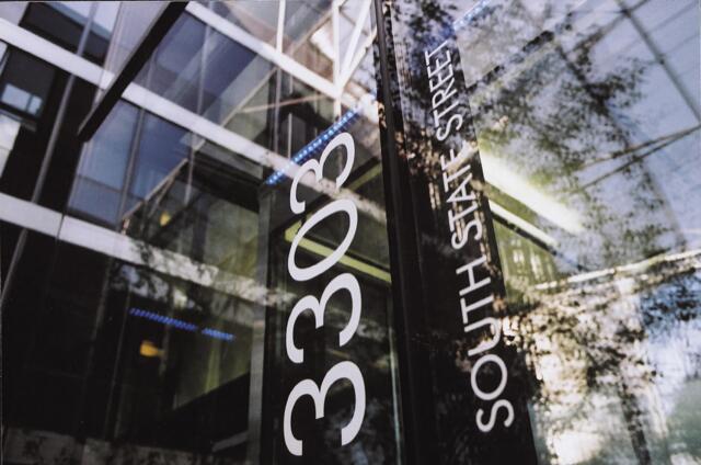 Glass building entrance with reflections of trees, showing the address "3303 South State Street" on the glass. Patterns of light and shadow create an abstract urban scene.
