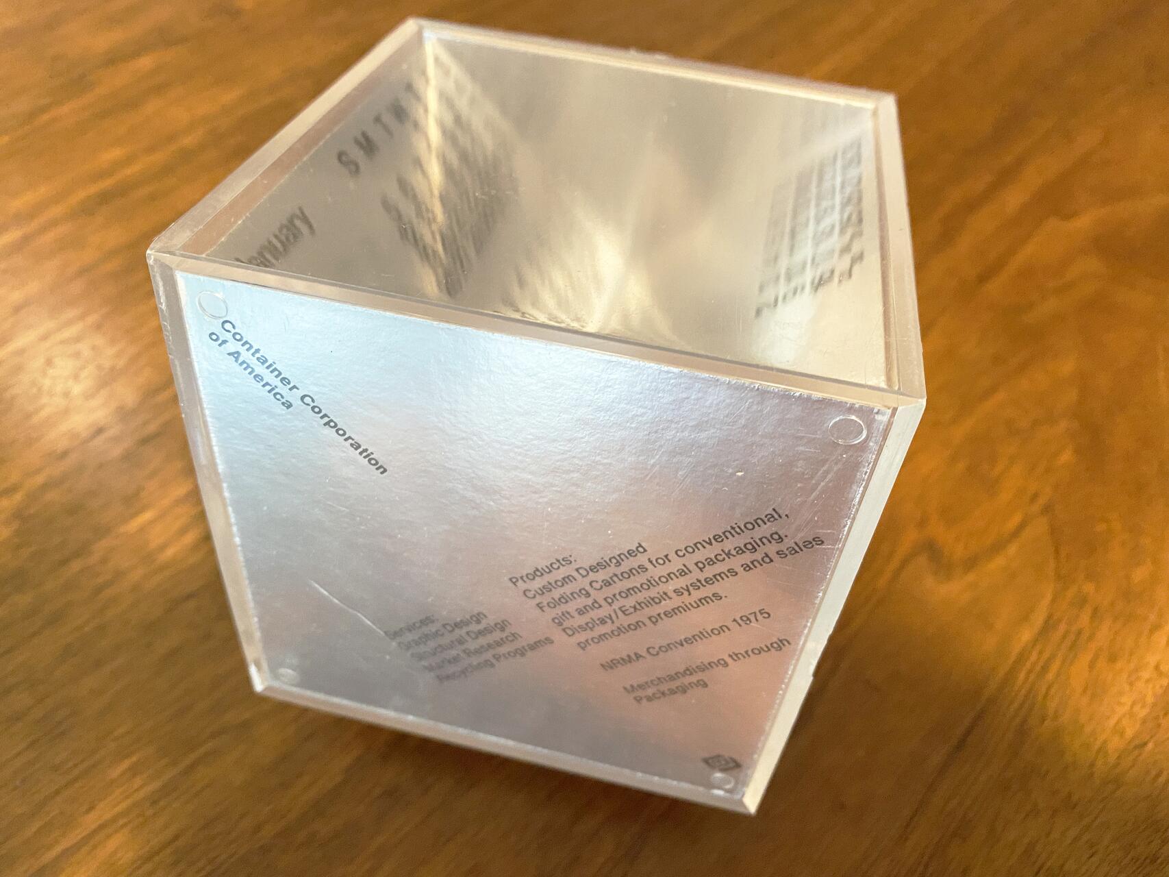Clear cube with text, including "Container Corporation of America," on a wooden surface.
