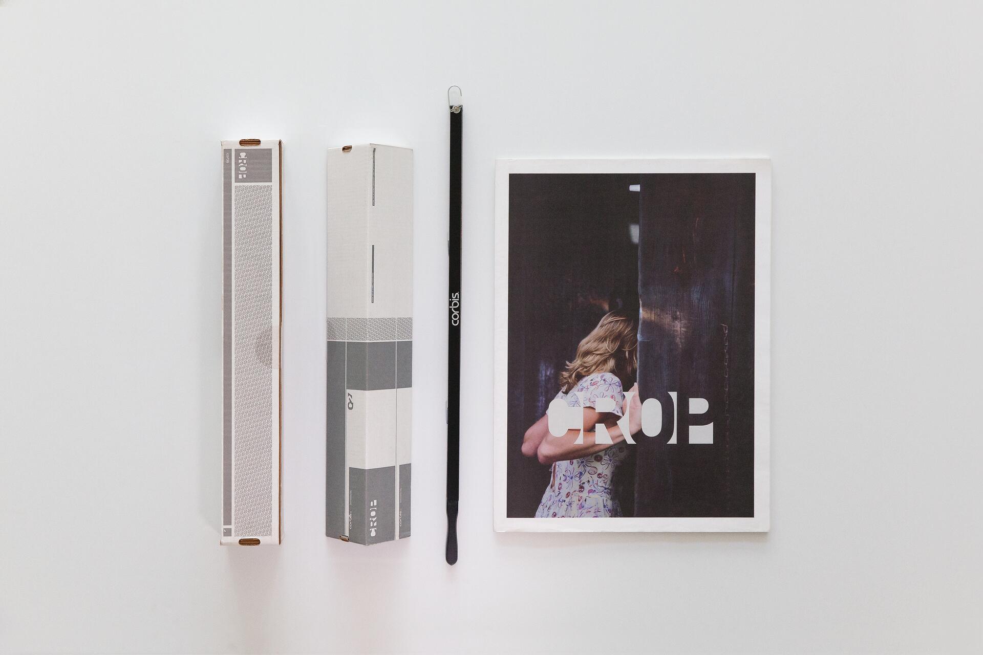 Two rectangular boxes, a long black tool labeled "corbis," and a print showing a woman peering through a gap in wooden planks with the word "CROP" overlaying her. The setting is minimal and clean.