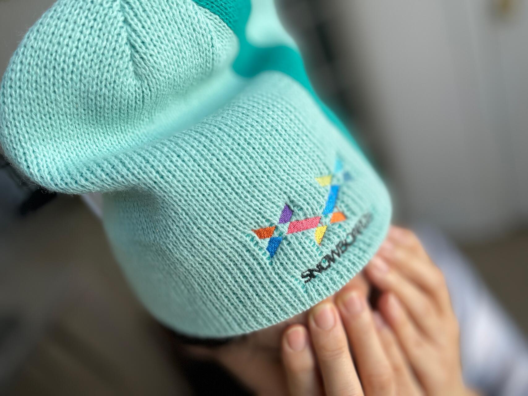 A person wearing a light blue beanie with colorful geometric embroidery partly covers their face with hands.