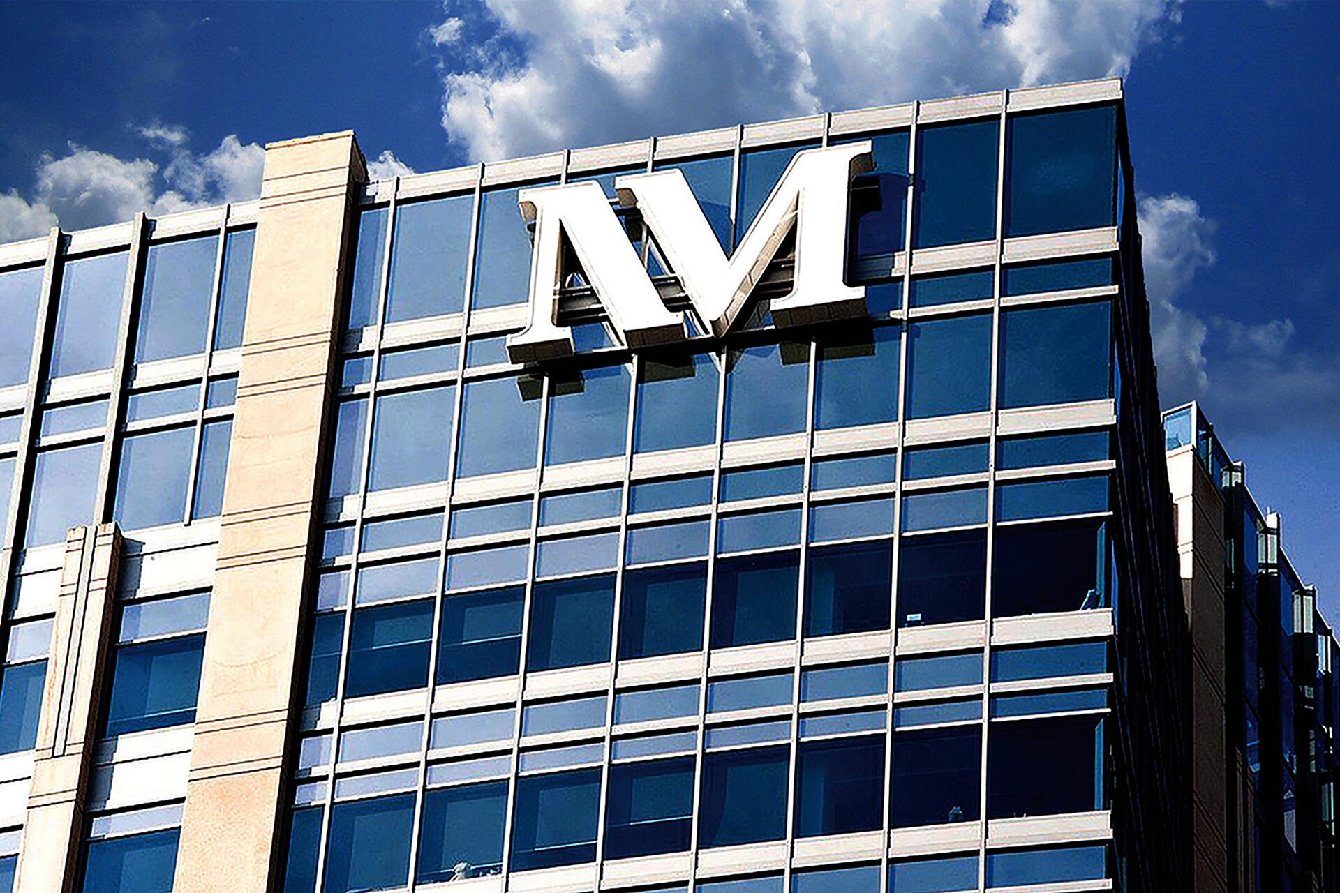 Glass office building with a large "M" logo on top, set against a blue sky with clouds.