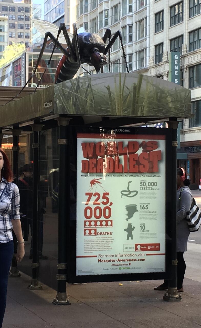 A giant mosquito sculpture on top of a city bus stop shelter, with a poster showing statistics about mosquito-related deaths. Two people walk past on the sidewalk. Tall buildings are visible in the background.