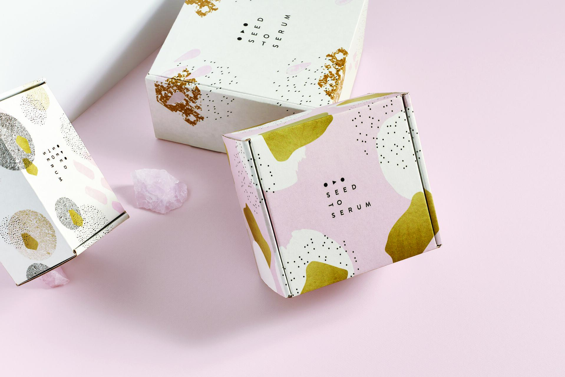 Colorful boxes with abstract patterns, labeled "Seed to Serum," on a pale pink surface, accompanied by rose quartz crystals.
