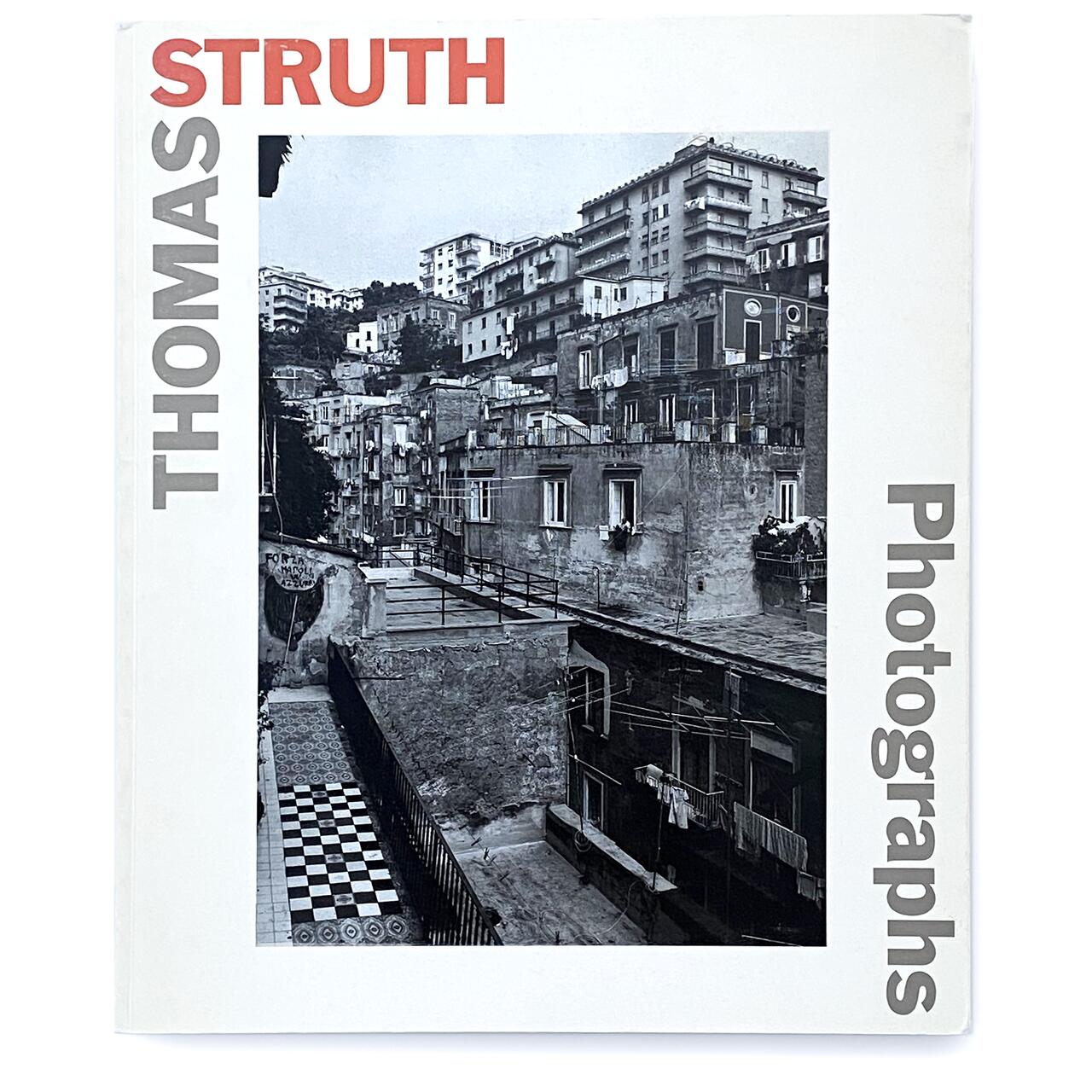 Black and white photo of an urban area with aged buildings, balconies, and a checkered courtyard. Book cover titled "Thomas Struth Photographs" with the title in red and black letters.