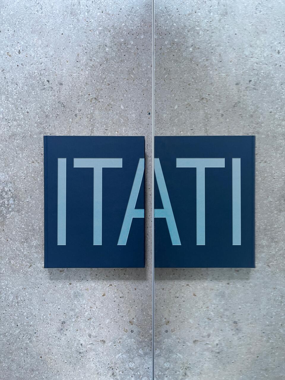 A dark blue book with large, bold letters "ITATI" reflecting against a shiny surface on a lightly textured stone background.