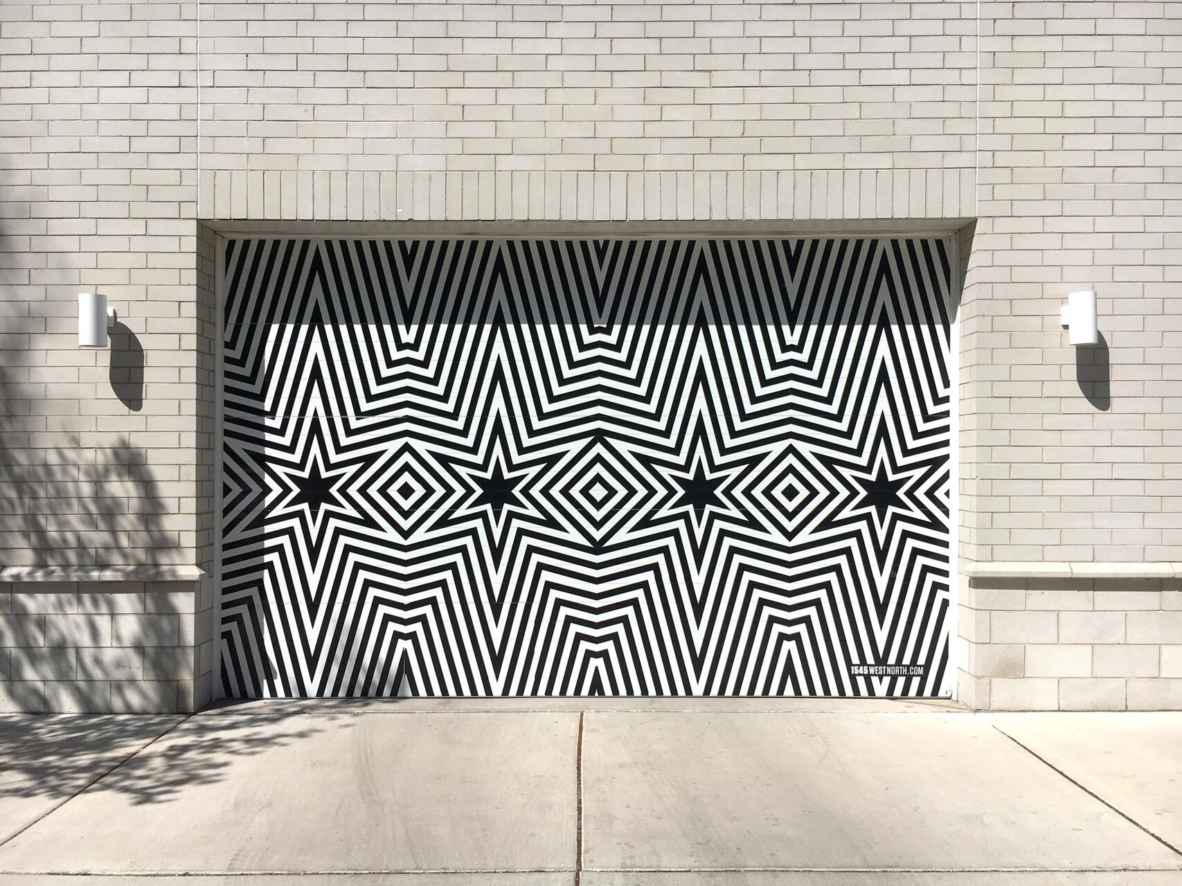 Garage door with black and white geometric star pattern on a light brick wall, with shadows cast from nearby trees.