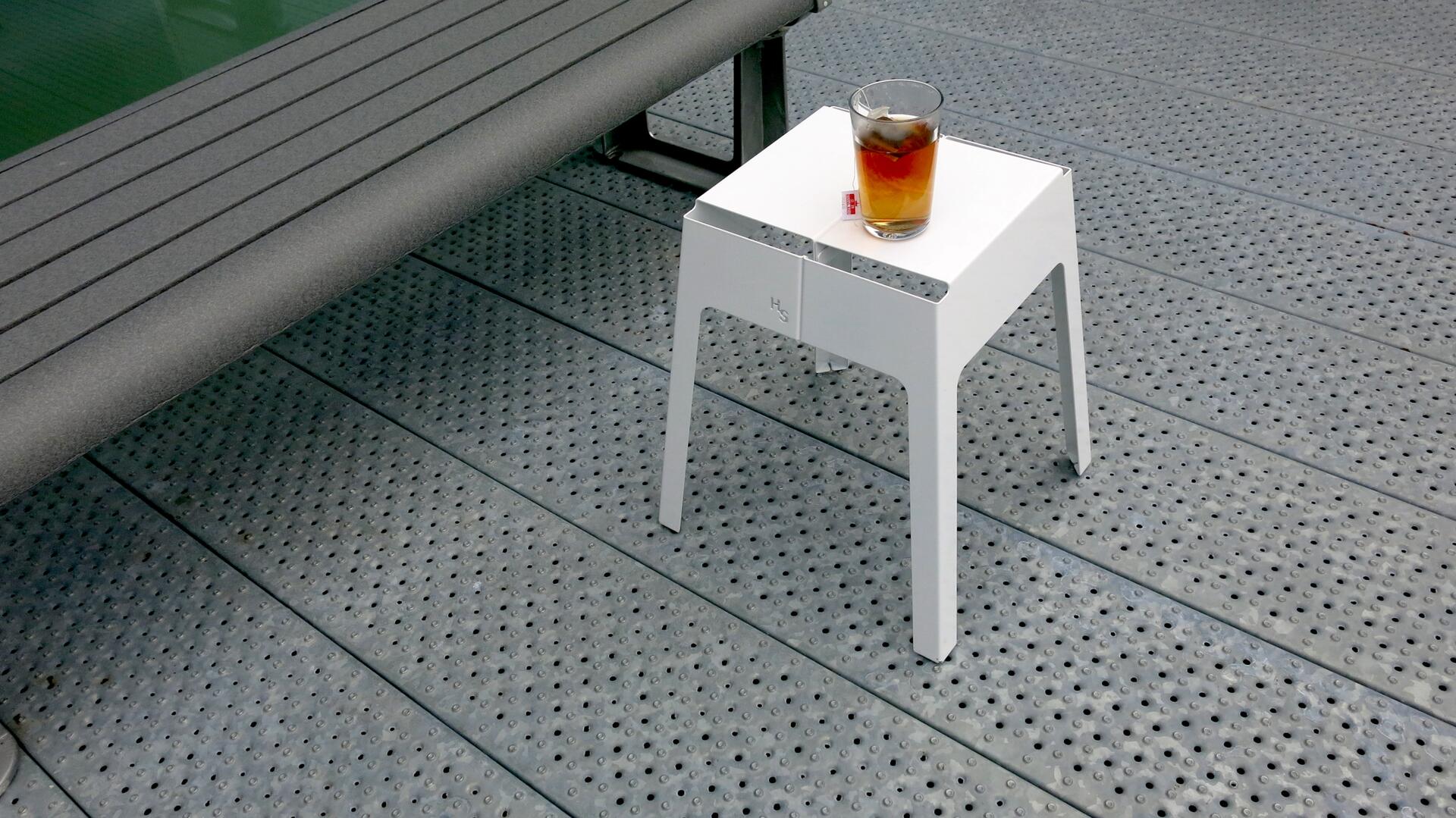 A glass of iced tea on a small white table, next to a gray bench, on a perforated metal deck.