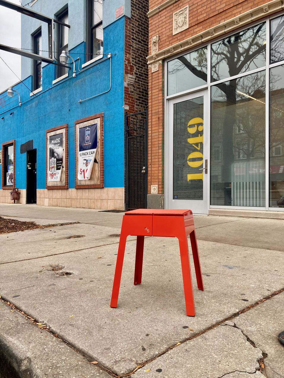 A bright red stool sits on a cracked sidewalk in an urban setting, with colorful blue and brick buildings in the background. Posters and large glass windows are visible, creating a lively city street vibe.