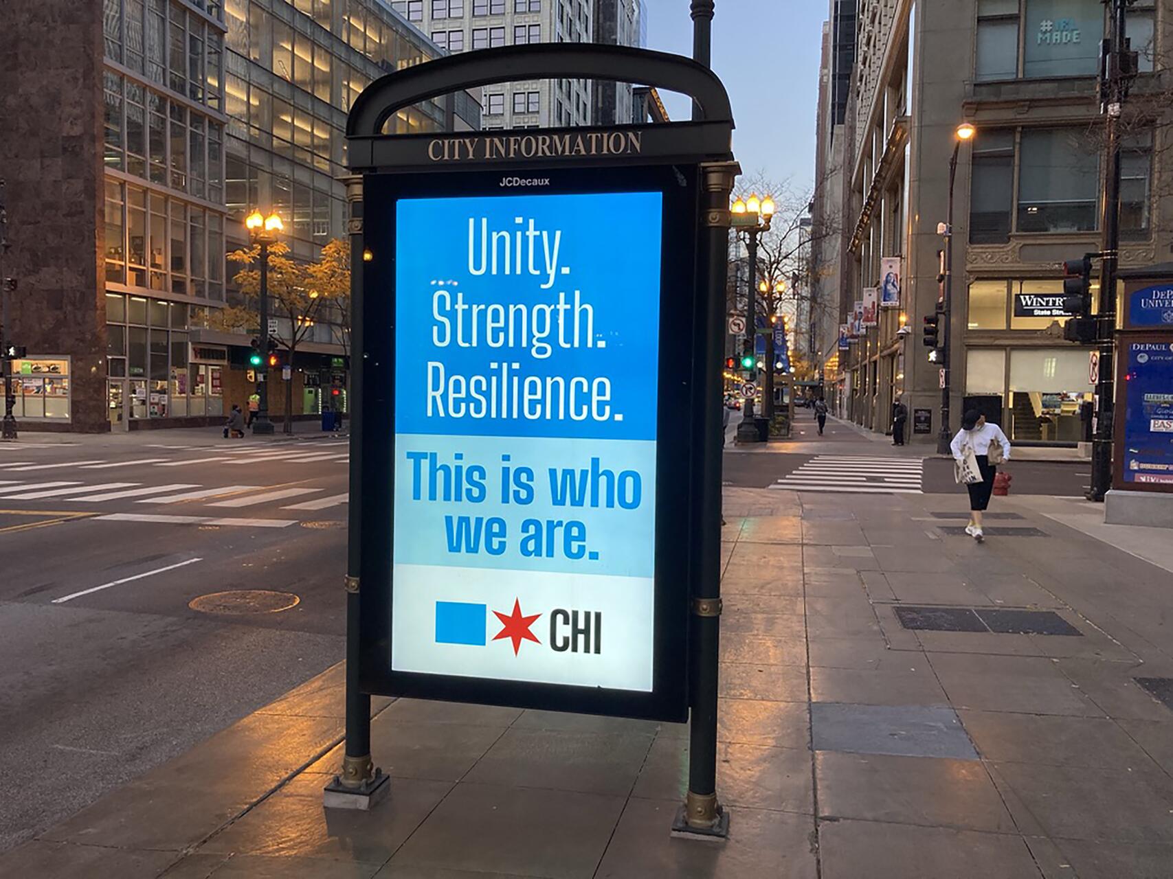 Street scene with a bus stop ad displaying a blue and white poster with the words "Unity. Strength. Resilience. This is who we are." The backdrop shows a city intersection and buildings illuminated by streetlights.